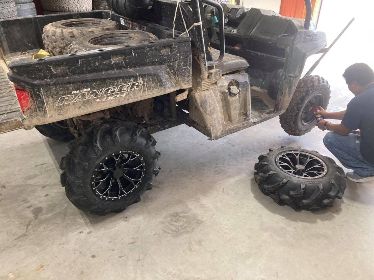 Utility vehicle with a flat tire in a garage, with a loose wheel on the floor and a man working nearby.