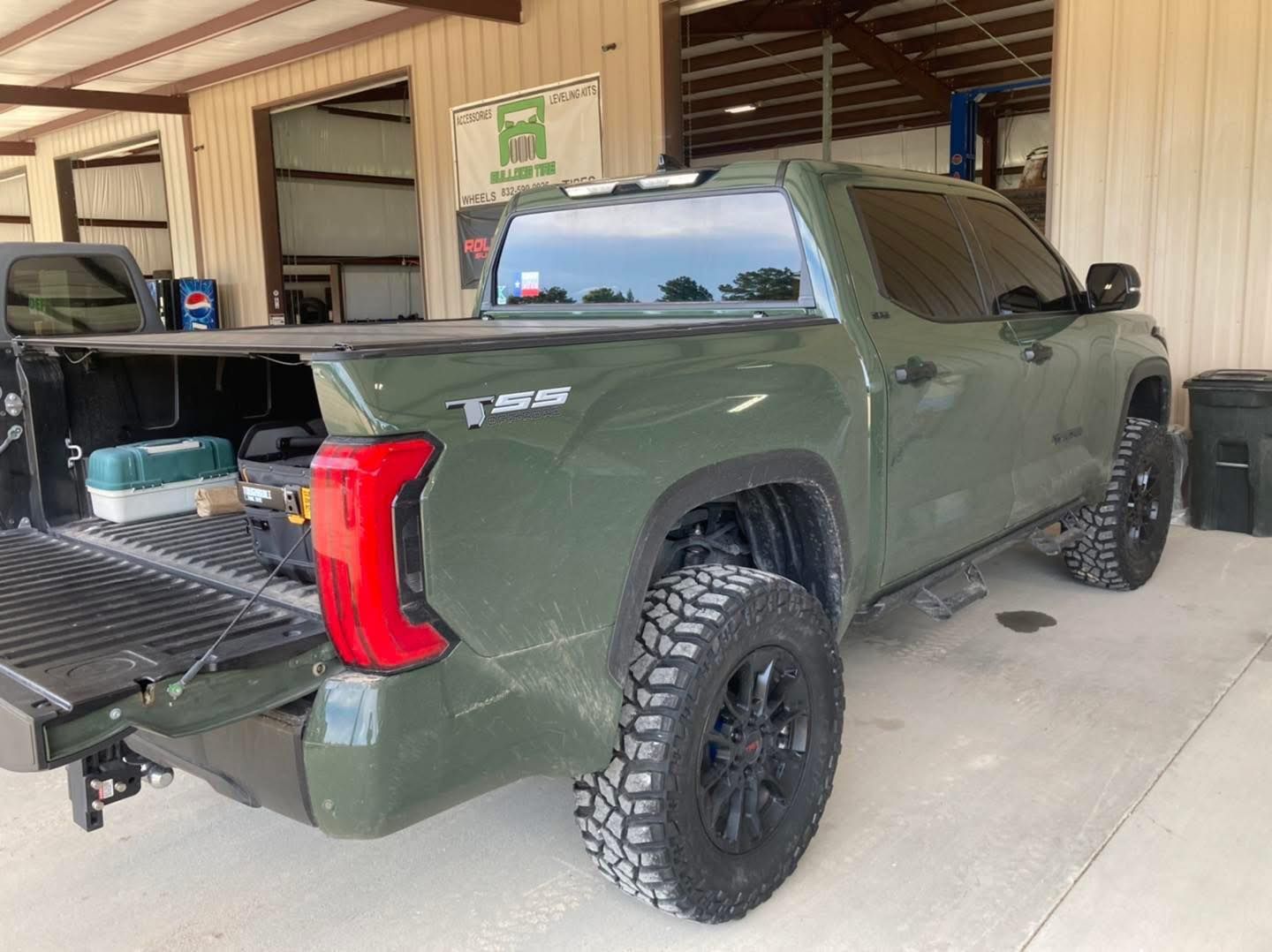 Green pickup truck parked in a garage with the tailgate open, showing the rear bed and off-road tires
