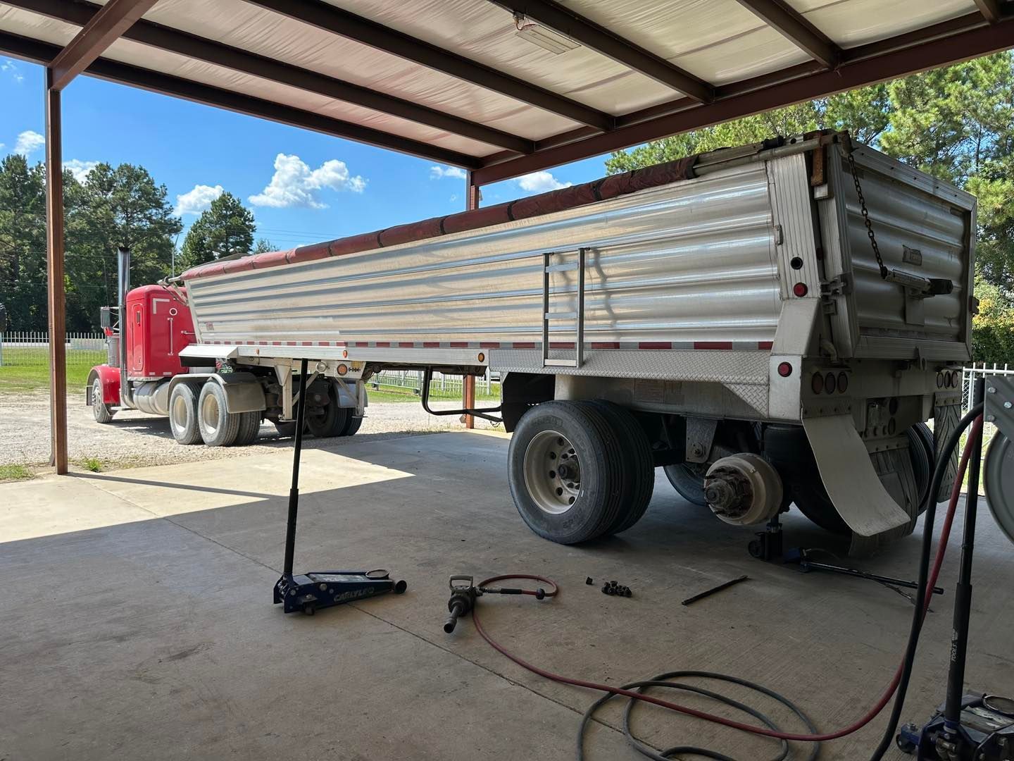 Red semi-truck with a silver dump trailer parked under a metal shed, tools and cables on the ground.