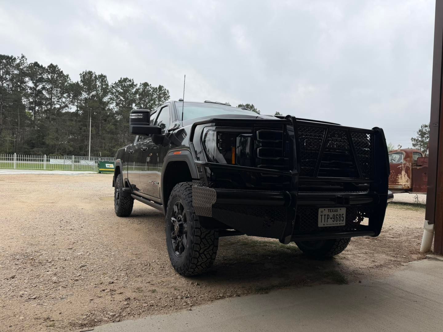 Black lifted pickup truck parked on gravel near a garage, viewed from the rear side.