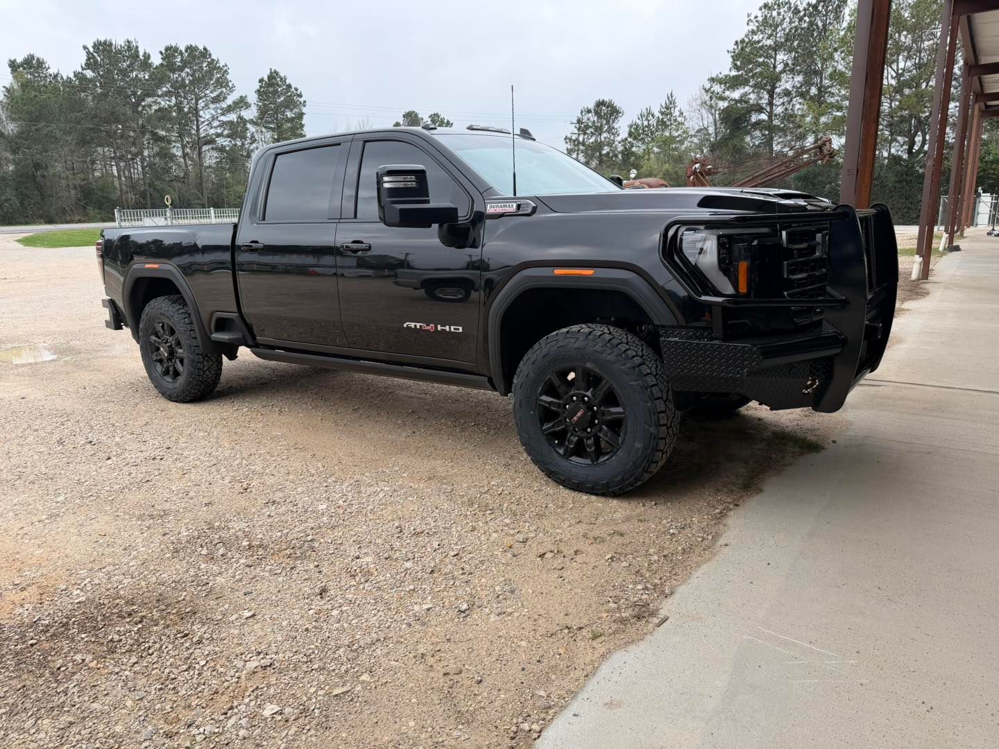 Black lifted pickup truck parked on a gravel lot beside a sidewalk, with trees in the background