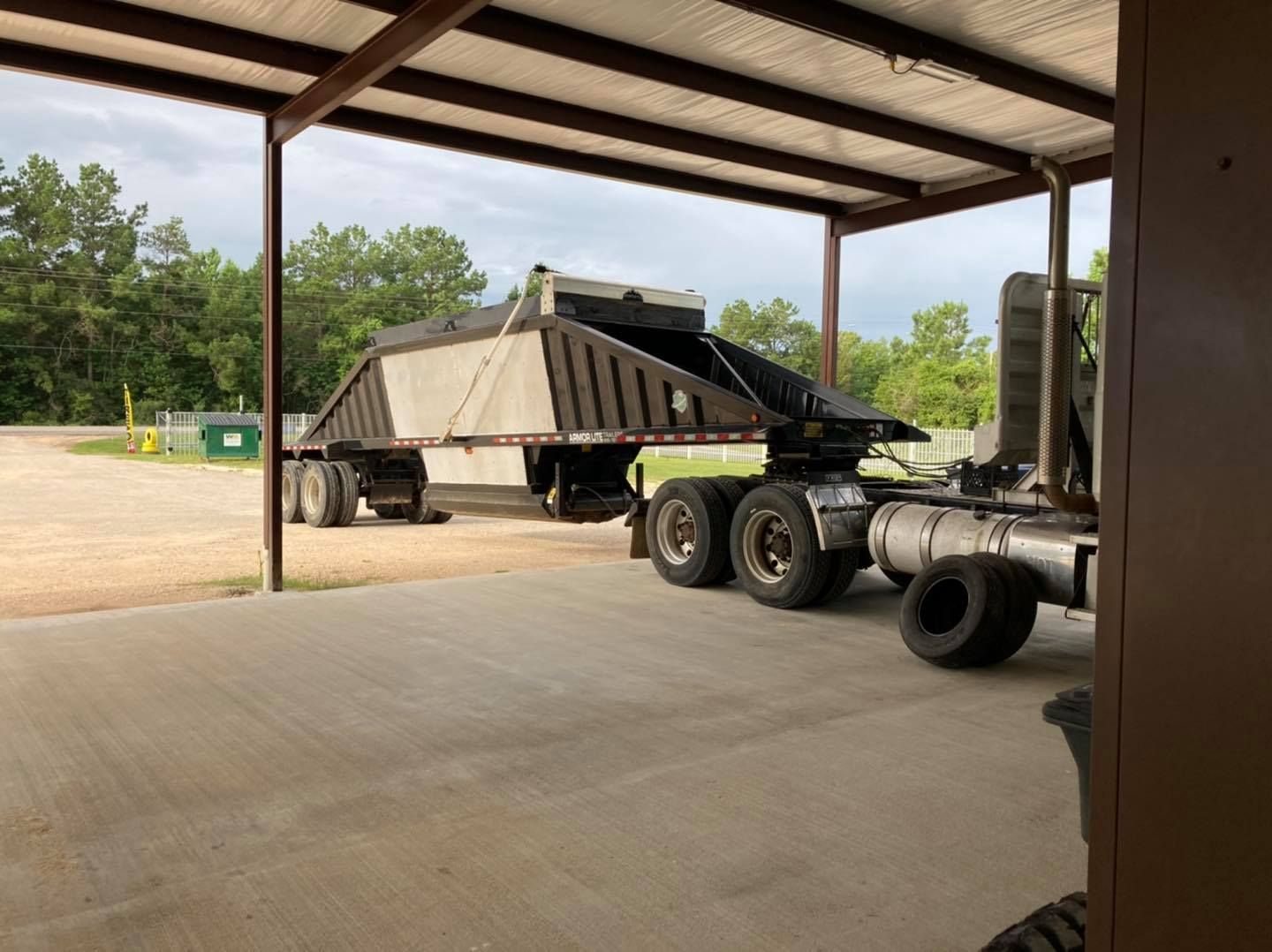Dump truck parked under a metal-roofed shed on a concrete pad, with trees and gravel outside.