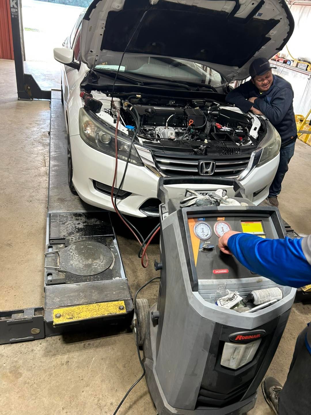 Mechanic charging a white Honda in a garage, with the hood open and a battery charger connected