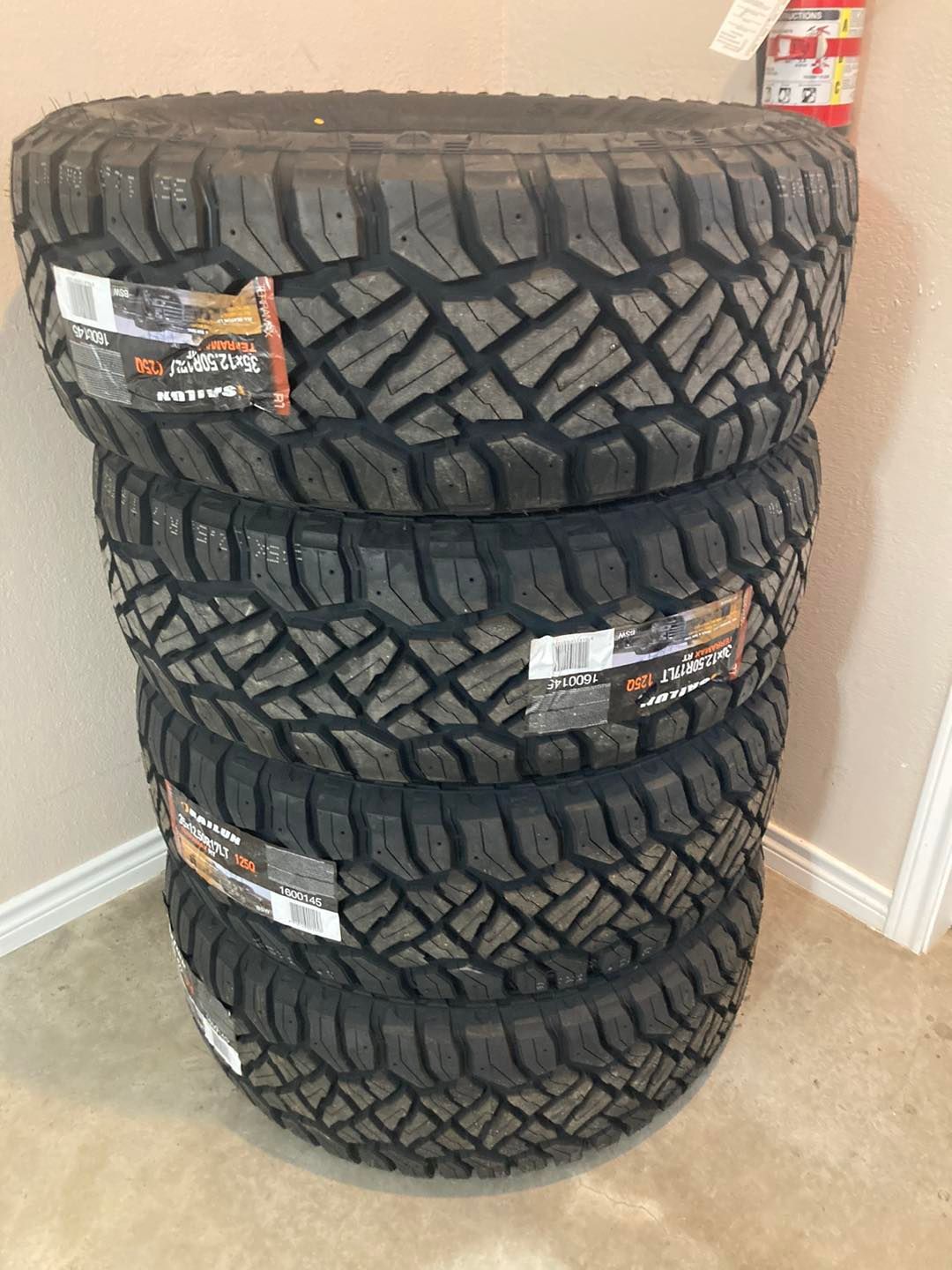 Stack of four off-road tires with deep tread patterns on a carpeted floor.