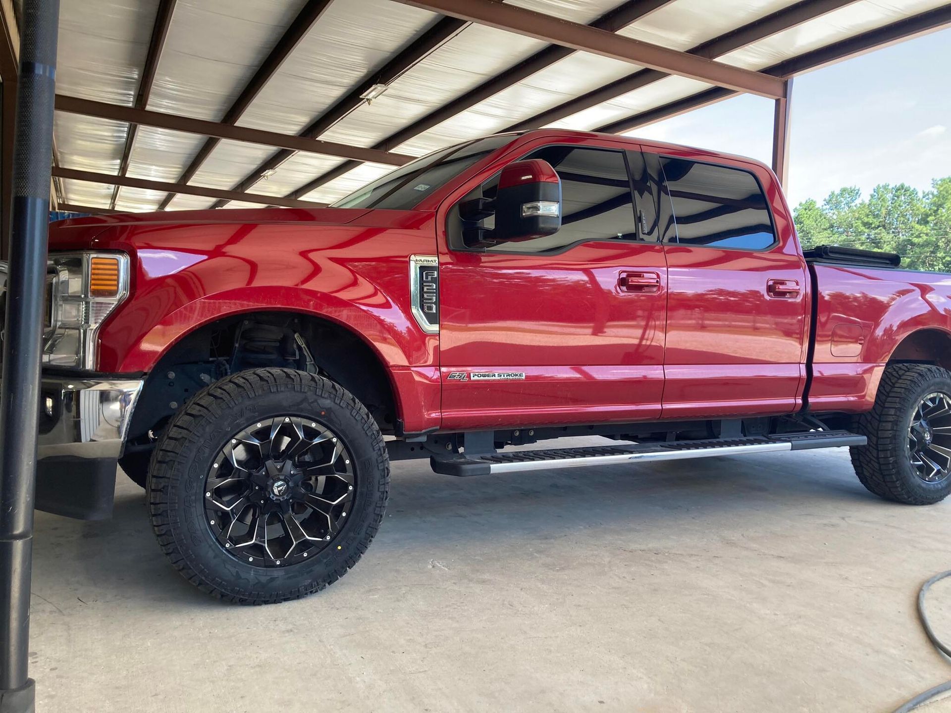 Red lifted pickup truck parked under a carport, side view with black off-road wheels.