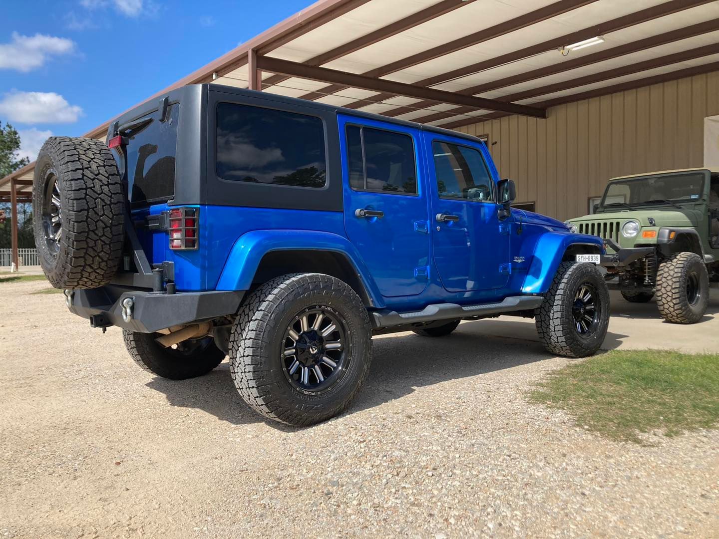 Blue Jeep Wrangler parked under a carport beside a green off-road vehicle.