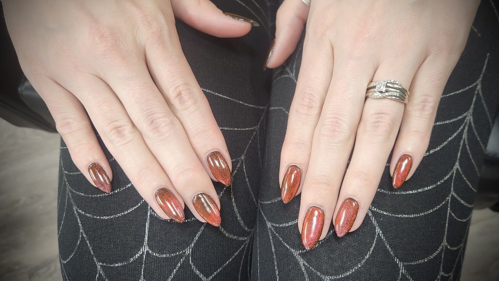 Hands with long, pointed nails painted with a brown and orange ombre design, resting on spiderweb-patterned fabric.