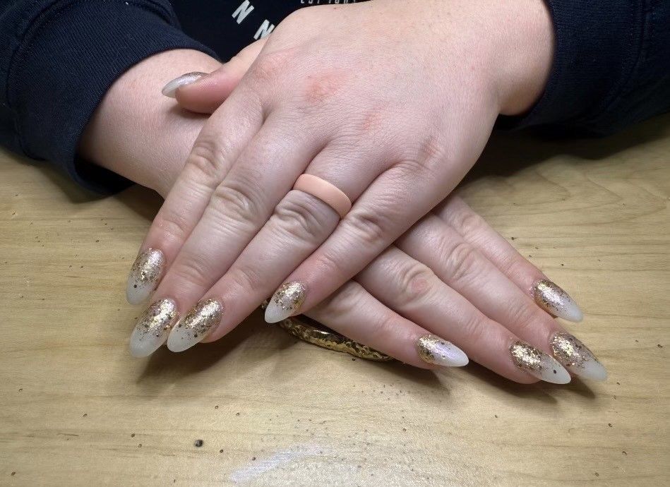 Hands with almond-shaped nails, gold glitter tips and a pink ring, crossed on a wood surface.