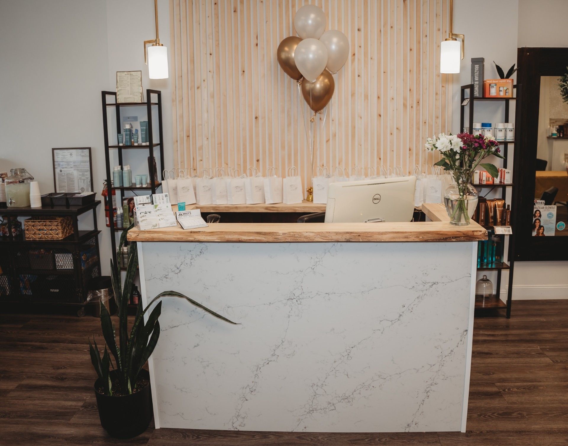 Reception desk in a beauty salon, with a wooden countertop and textured white facade, wooden wall backdrop, balloons, and shelving.