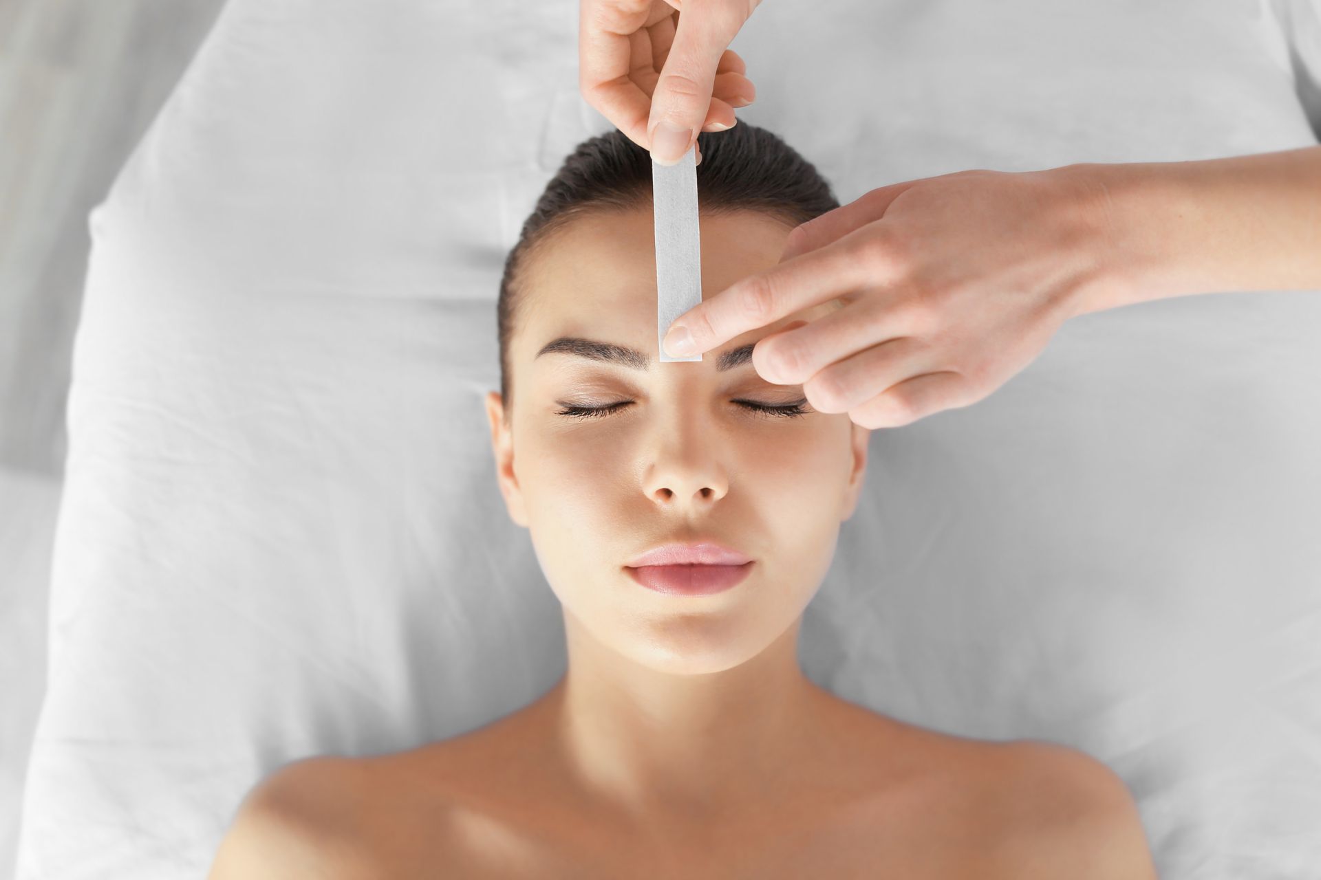 Woman having her brow waxed, eyes closed, with a white wax strip on her forehead.