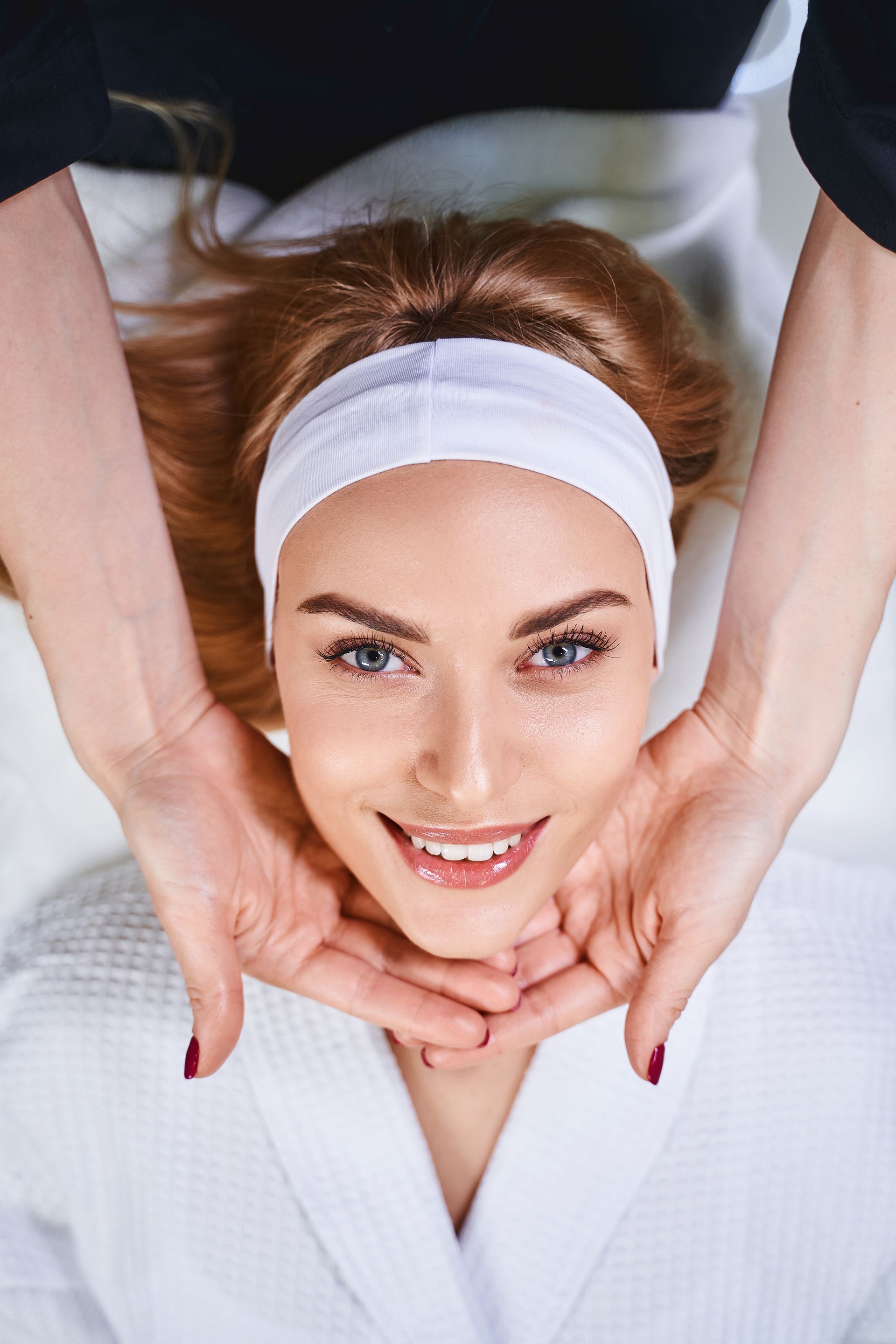 Woman receiving spa treatment; hands cradling face, smiling, wearing headband, white robe.