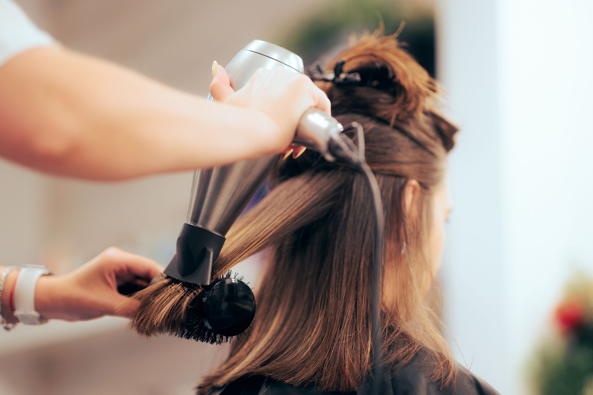 Hair stylist using a blow dryer and round brush on a client's hair in a salon.