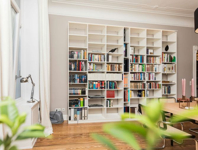 Bright living room with white bookshelves filled with books, wooden floor, and blurred green plants in foreground