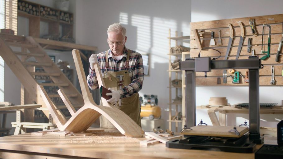 Man working in a bright workshop, shaping a wooden chair at a workbench with tools nearby