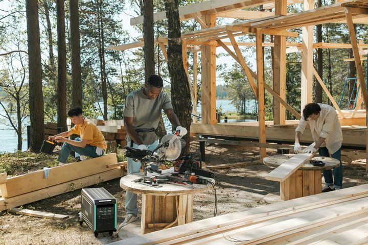 Three people doing carpentry on a wooden deck at a forested lakeside cabin site.