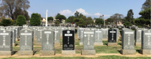 Rows of tombstones in a cemetery with a dark black headstone centered in the middle. Trees and blue sky in the background.