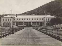 A long, light-colored building with a bridge leading to it. Flags fly on poles. Mountain in the background.
