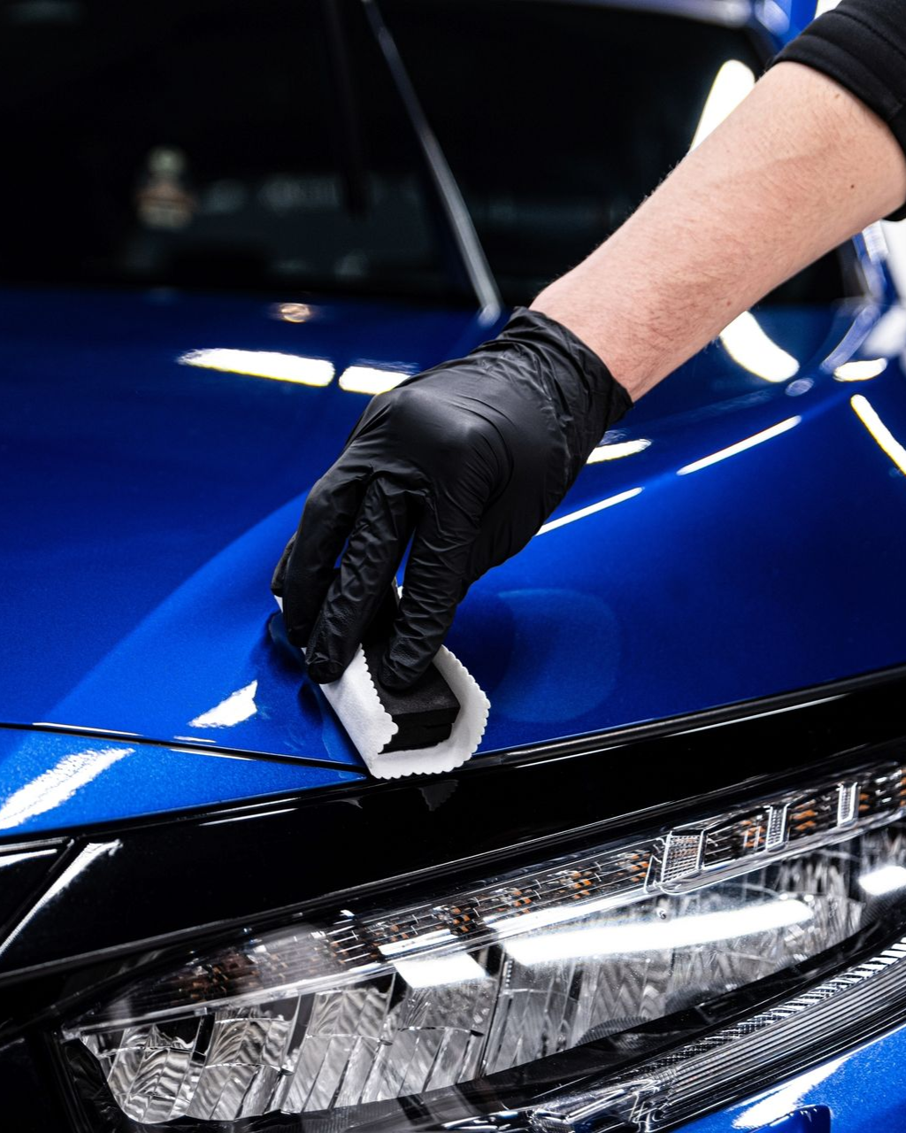 Gloved hand applying ceramic coating to a blue car's hood with a white applicator.