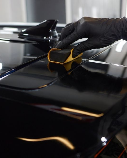 Gloved hand applying a ceramic coating to a black car surface with a yellow applicator.