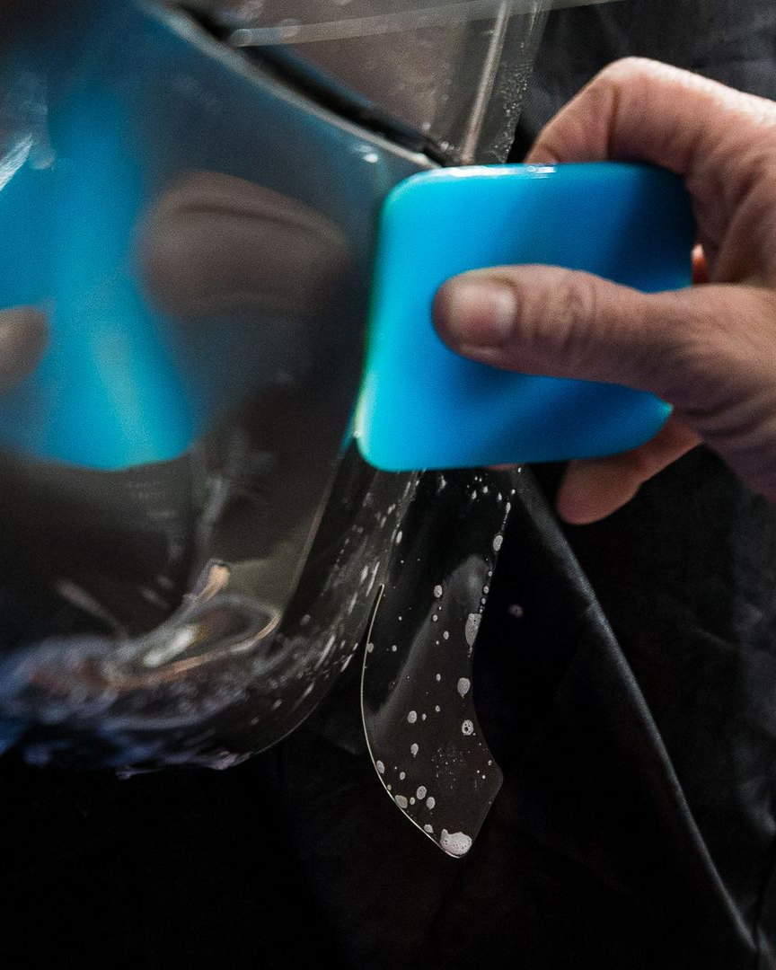 Hand using a blue squeegee to apply clear film to a car surface. Film has soapy water.