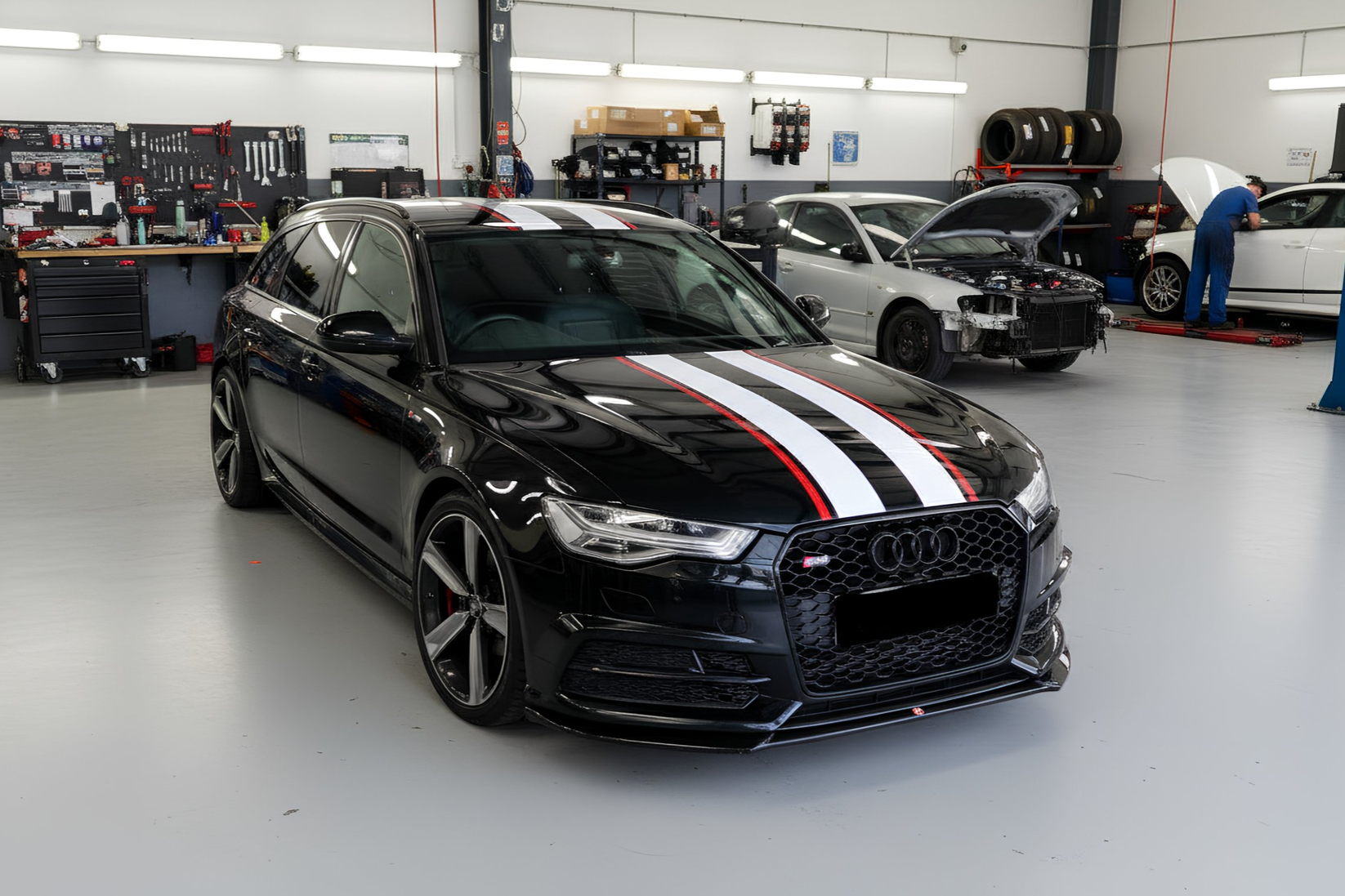 Black Audi with white and red stripes in a garage; other cars and mechanics visible.