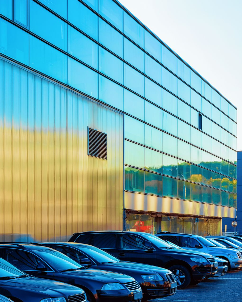 Cars parked in front of a modern building with a glass facade, reflecting the sky.