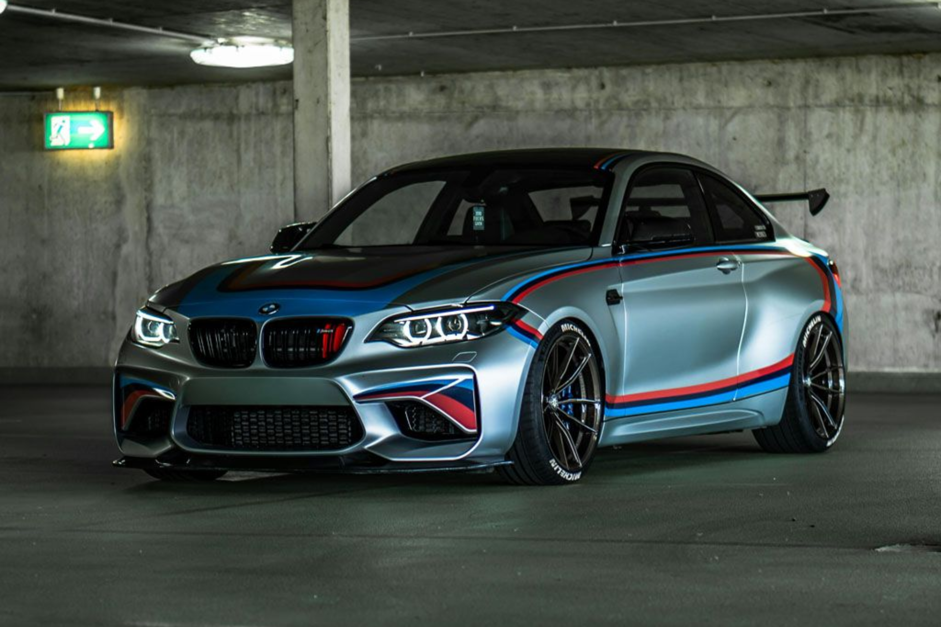 Silver BMW M2 sports car with racing stripes parked in a parking garage.