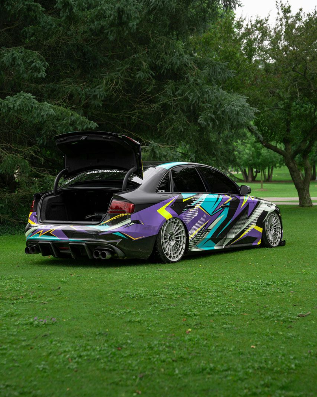 Black car with graphic wrap, trunk open, on grass.