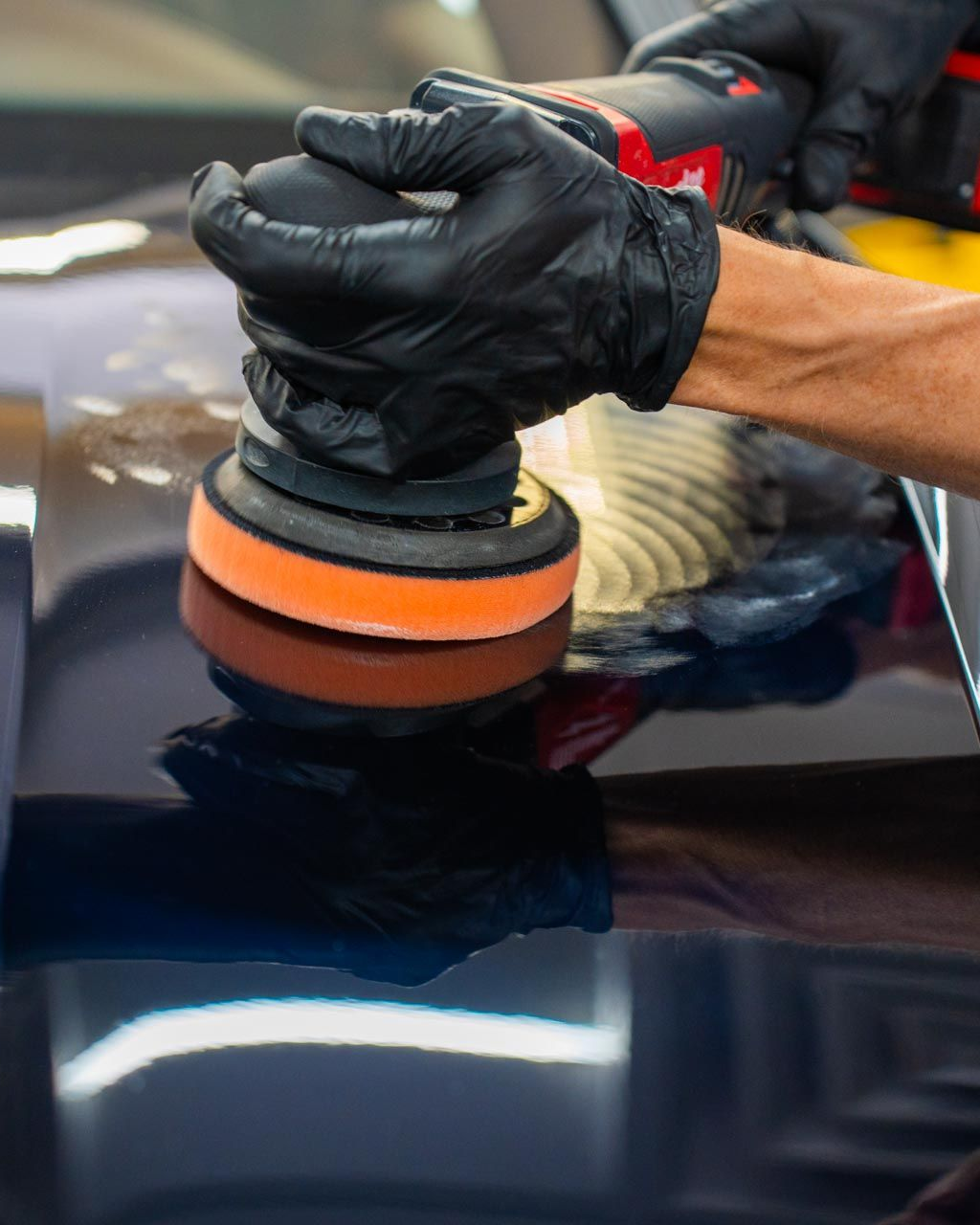 Person polishing a car's dark blue surface with a powered buffer; wearing black gloves.