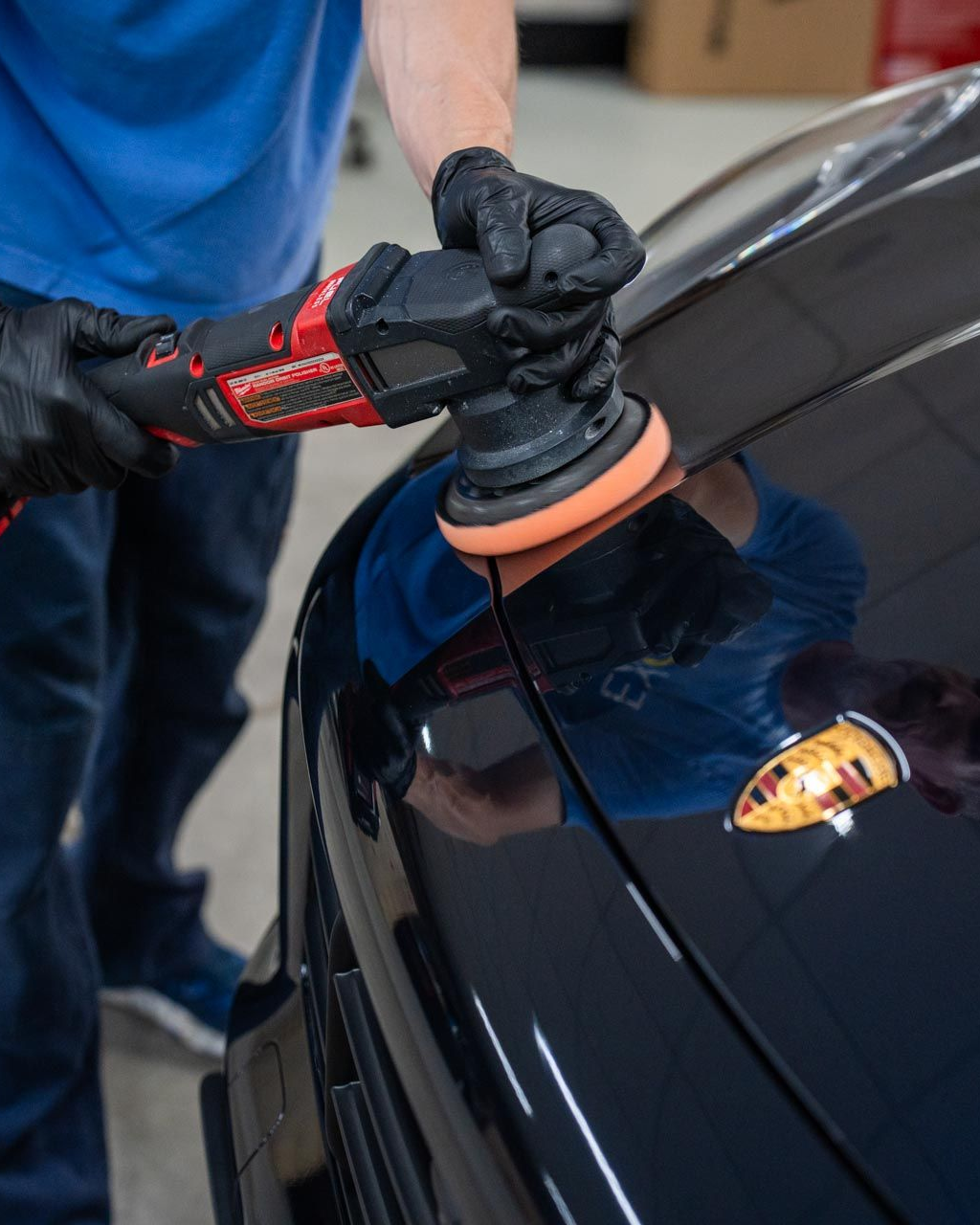 Person polishing a black car with a rotary buffer, wearing gloves in a well-lit shop.