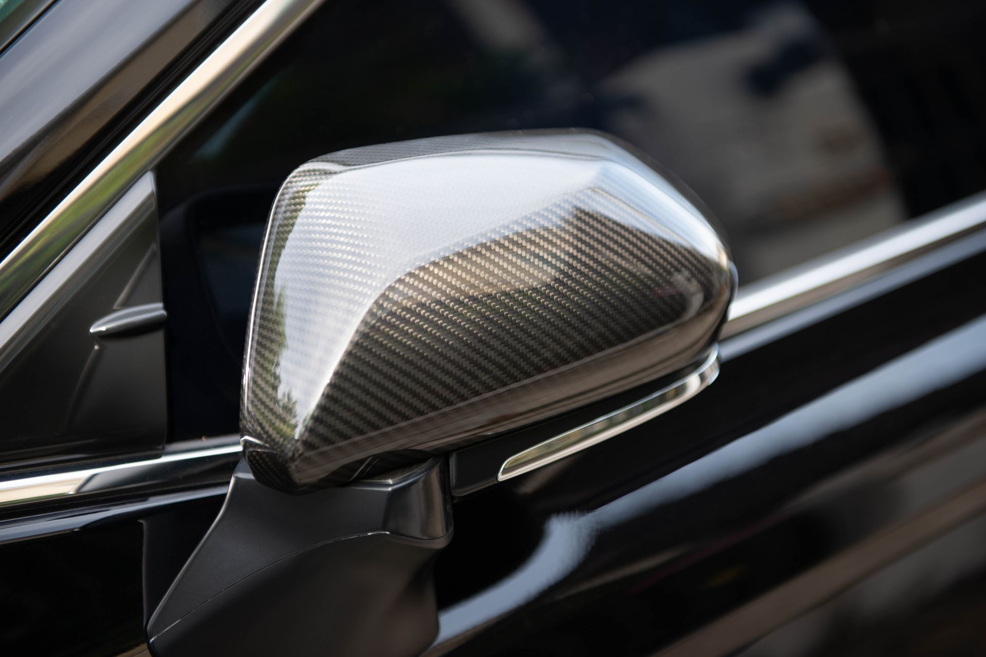 Carbon fiber car side mirror reflecting surroundings, mounted on a black vehicle.