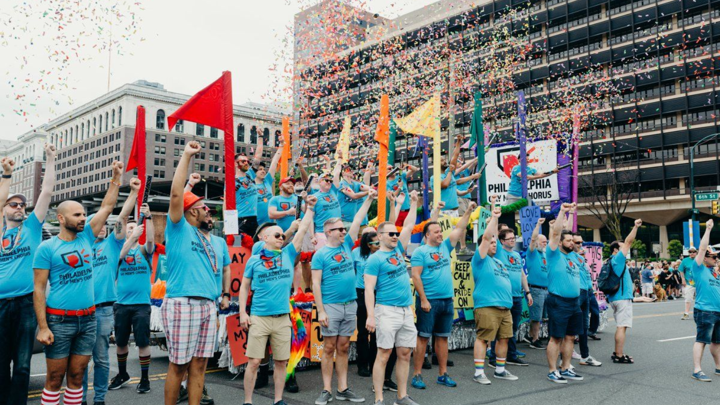 The Philadelphia Gay Men's Chorus (PGMC) in a street pride parade, with a rainbow flag and people cheering on both sides