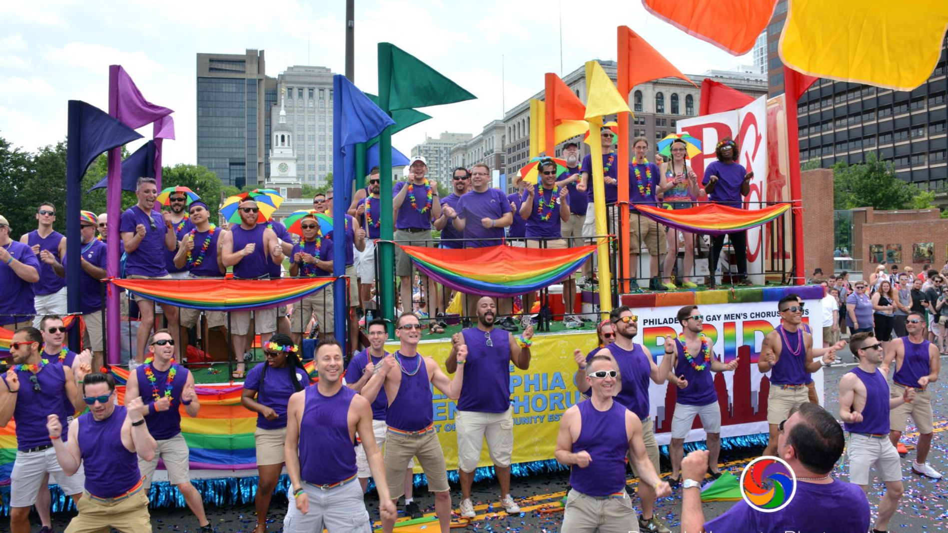 The Philadelphia Gay Men's Chorus (PGMC) in a street pride parade, with a rainbow flag and people cheering on both sides along Independence Mall in Old City.
