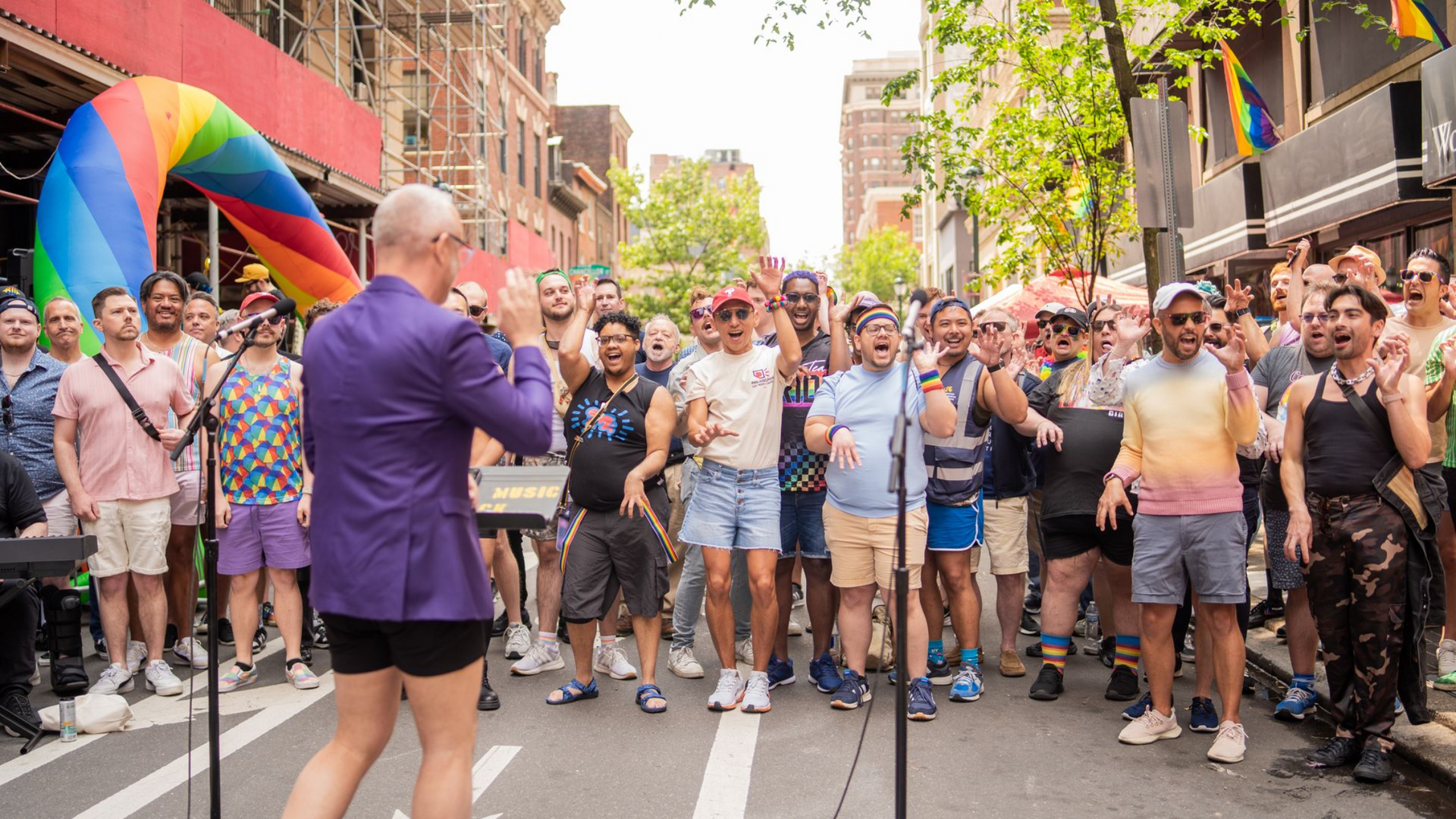 The Philadelphia Gay Men's Chorus (PGMC) in a street pride parade, with a rainbow flag and people cheering on both sides, dancing with raised hands to Lady Gaga's Bad Romance.
