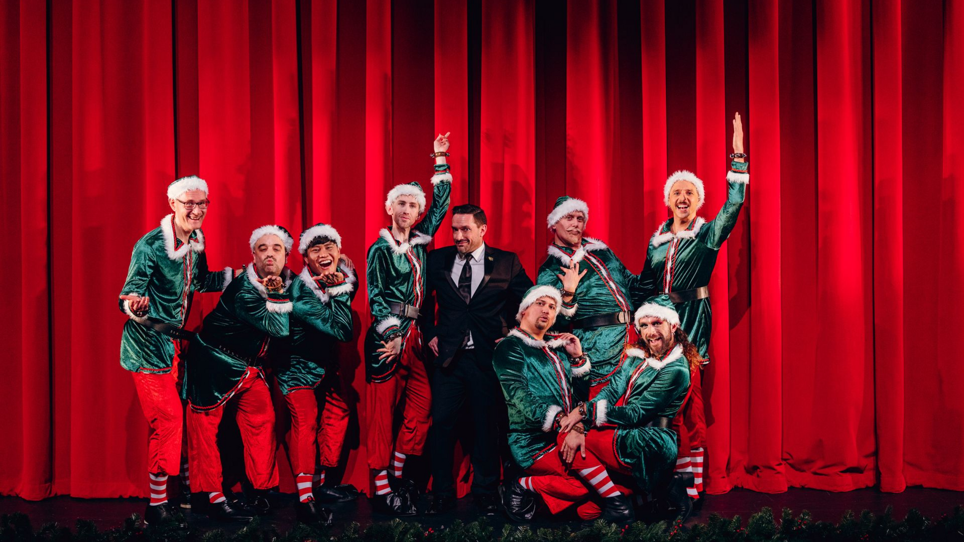 The Philadelphia Gay Men's Chorus, dressed in green and red elf costumes pose onstage before red curtains.