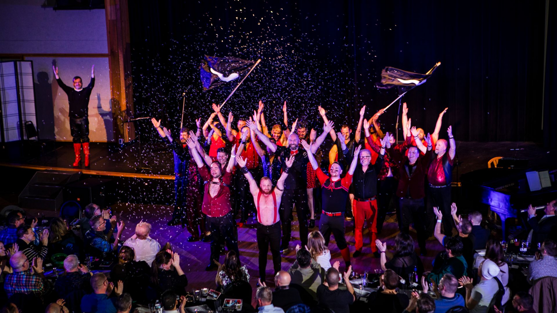 The Philadelphia Gay Men's Chorus (PGMC) performs on a stage with arms raised, confetti falling, and an audience watching below.