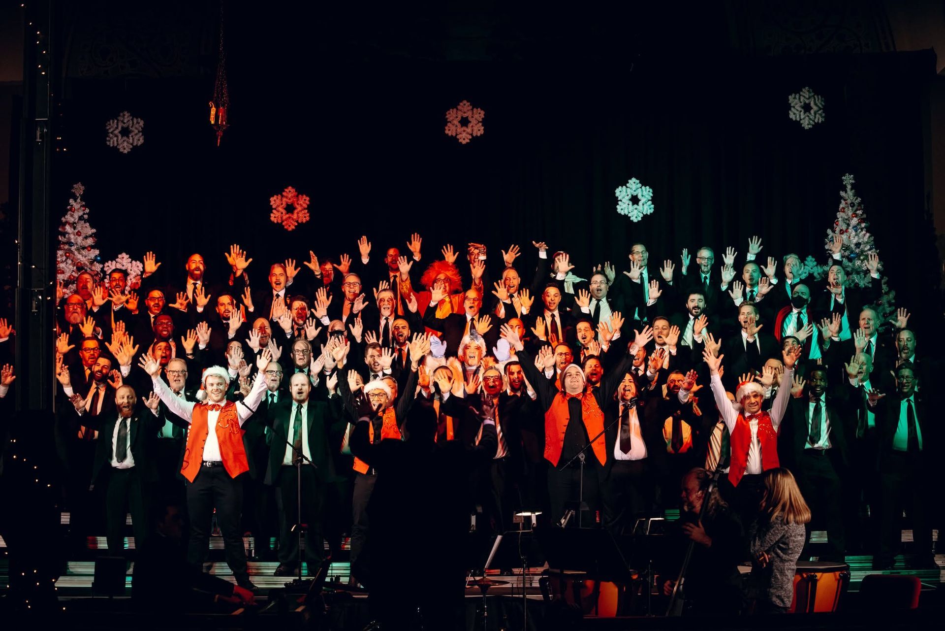 The Philadelphia Gay Men's Chorus (PGMC) is performing on a dark stage with red and green holiday decorations and snowflake lights