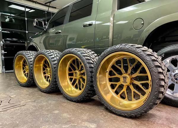 Four gold-rimmed truck wheels with large tires in front of a green pickup truck in a shop.