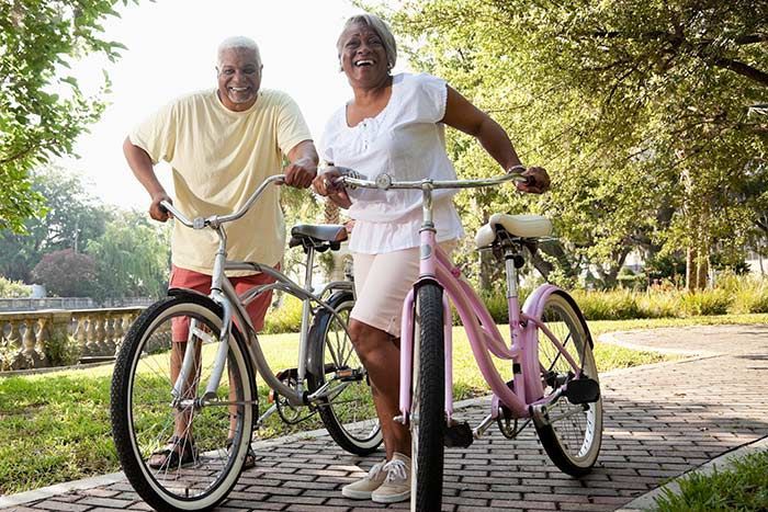 Two cyclists standing with bikes on a sunny park path, smiling beside trees.