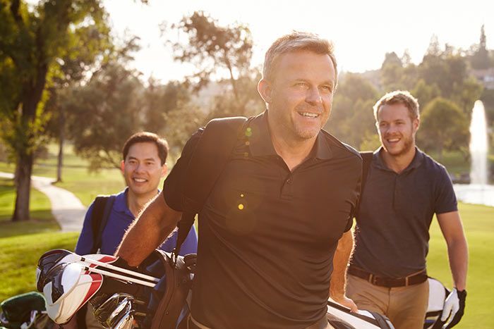 Three smiling golfers walking with clubs on a sunny course near a golf cart