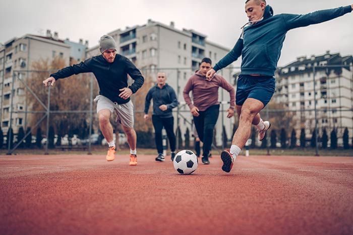 Four people playing soccer on an outdoor court, with one player kicking the ball toward the camera.