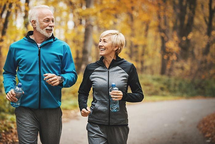 Two adults jogging on a tree-lined path in autumn, smiling and carrying water bottles.