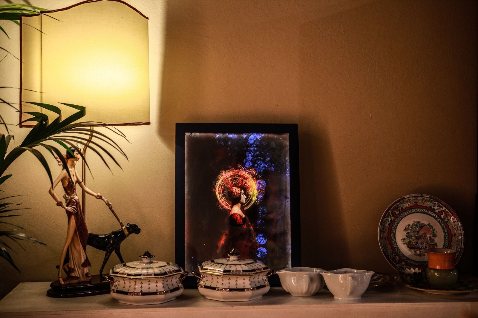 Un tavolo con una lampada, una statuetta, un ritratto illuminato incorniciato, due piatti con coperchio, due tazze e un piatto decorativo.