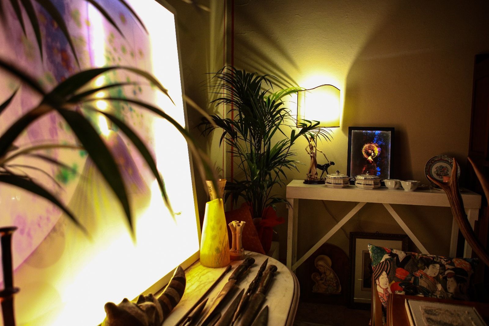 Una stanza calda e in penombra presenta una consolle con oggetti decorativi, una lampada e piante da interno vicino a un'installazione luminosa suggestiva.