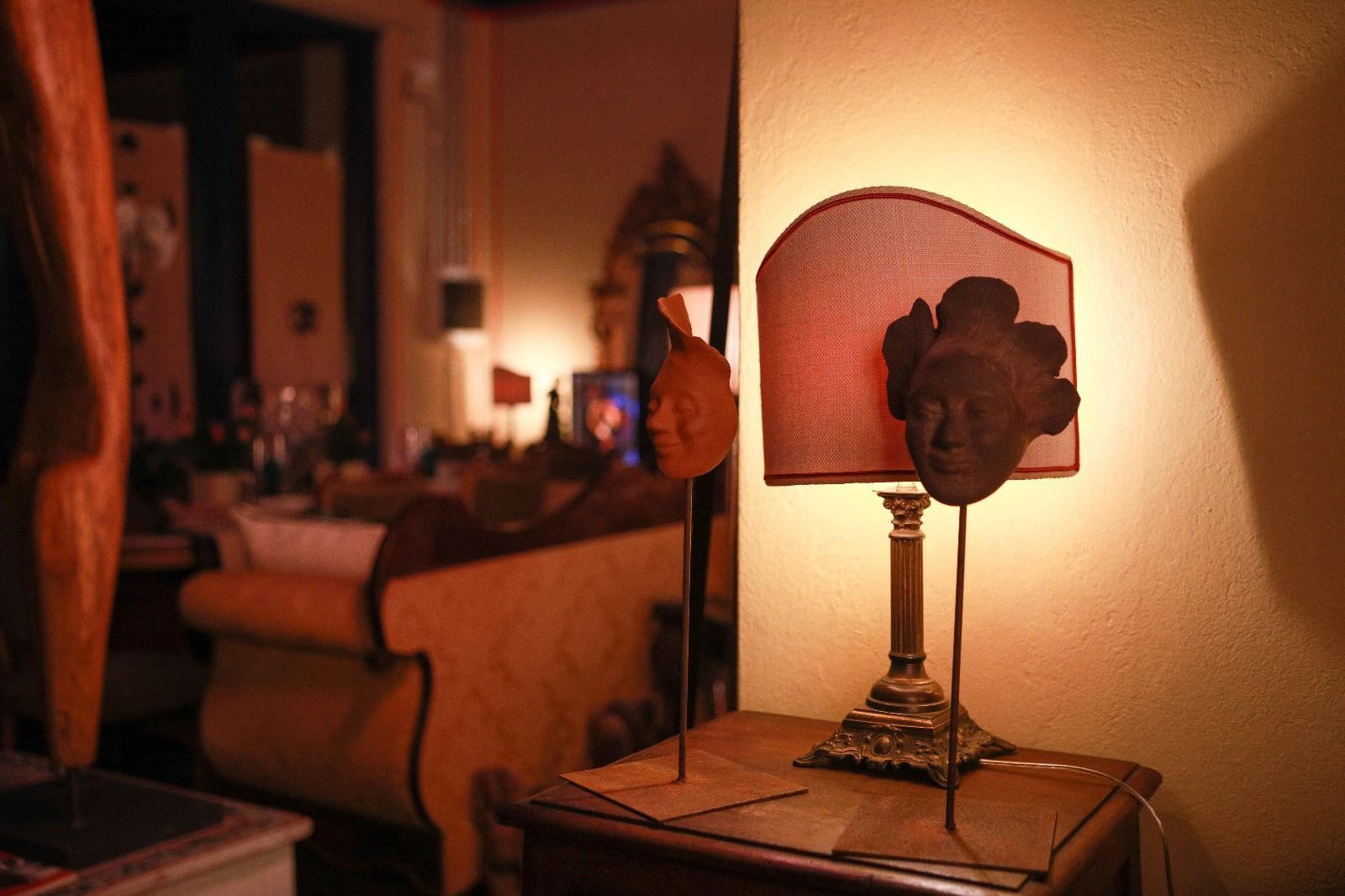 Una lampada con un paralume in tessuto rossastro è appoggiata su un tavolino di legno, affiancata da due sculture di teste su piedistalli di metallo.