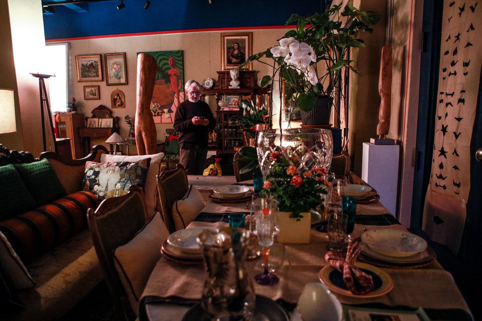 Una sala da pranzo illuminata da una luce calda, con un tavolo apparecchiato, piante da interno, pareti decorate con opere d'arte e una persona in piedi sullo sfondo.