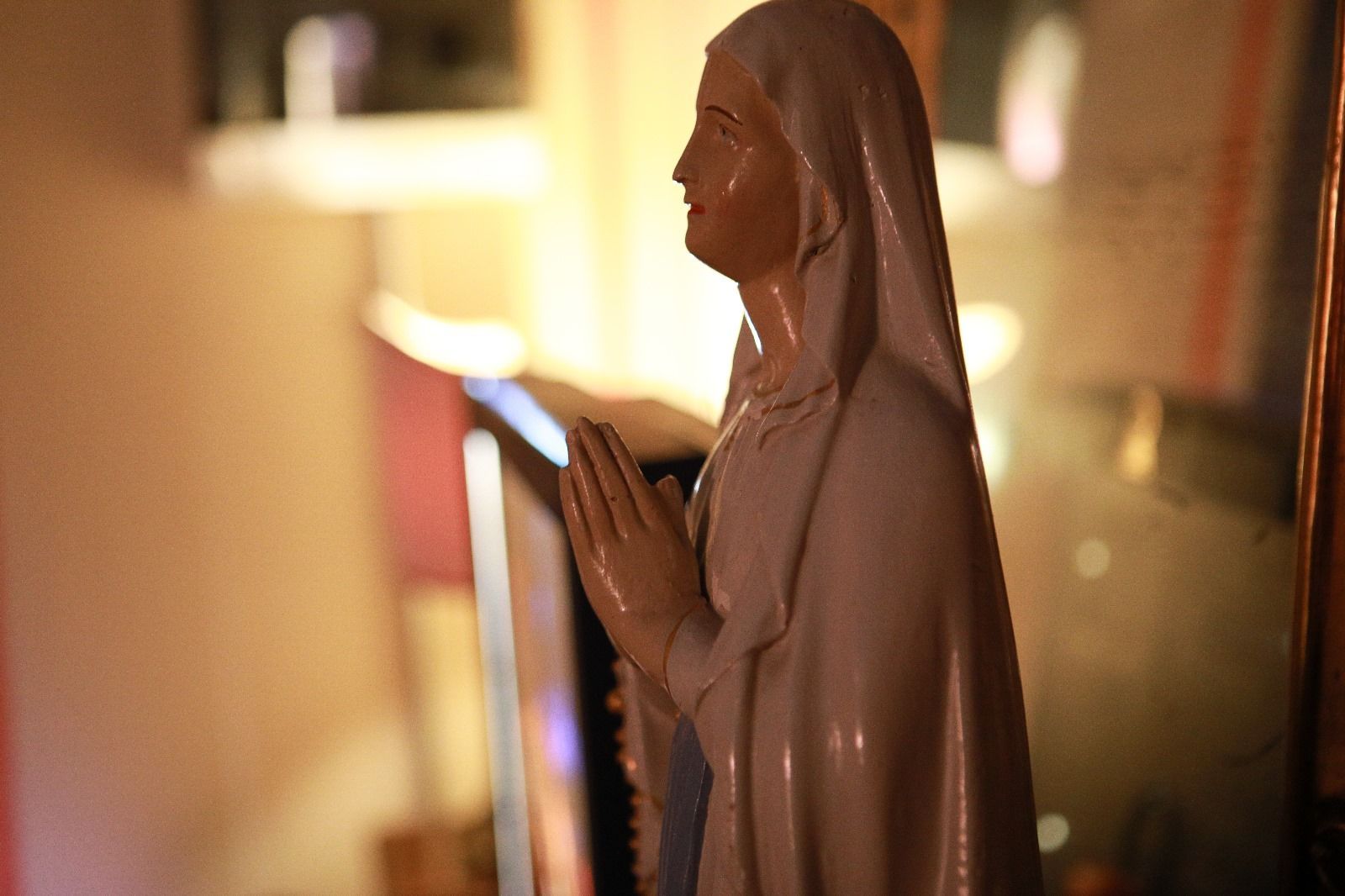 Una statua liscia e dai colori chiari raffigurante una figura con le mani giunte in preghiera, su uno sfondo leggermente sfocato.