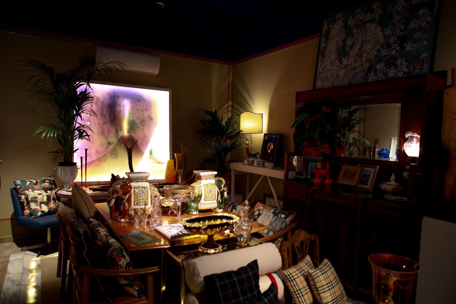 Una sala da pranzo eclettica e scarsamente illuminata, con un grande tavolo, vasi decorativi, rigogliose piante d'appartamento e mobili scuri.