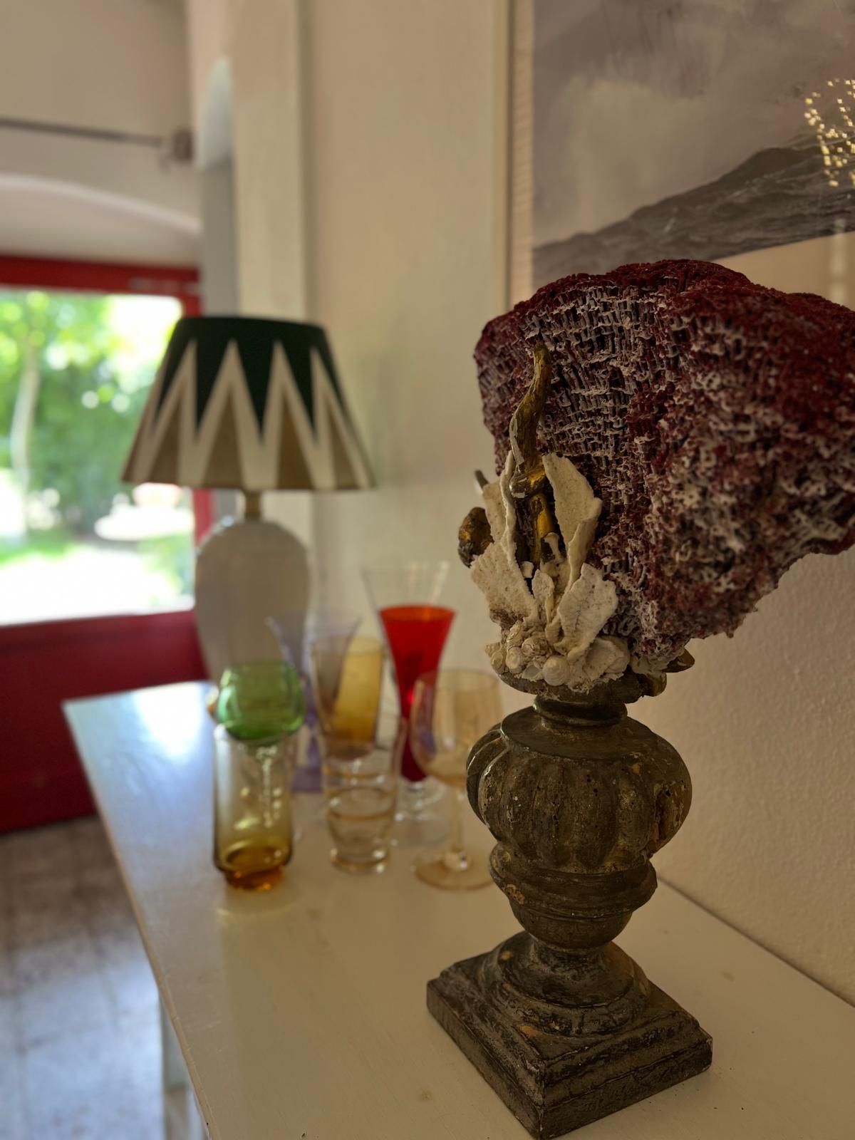 Un vaso decorativo con piedistallo in stile antico, posto su una mensola bianca vicino a una lampada decorata, contiene un pezzo di corallo rosso essiccato.