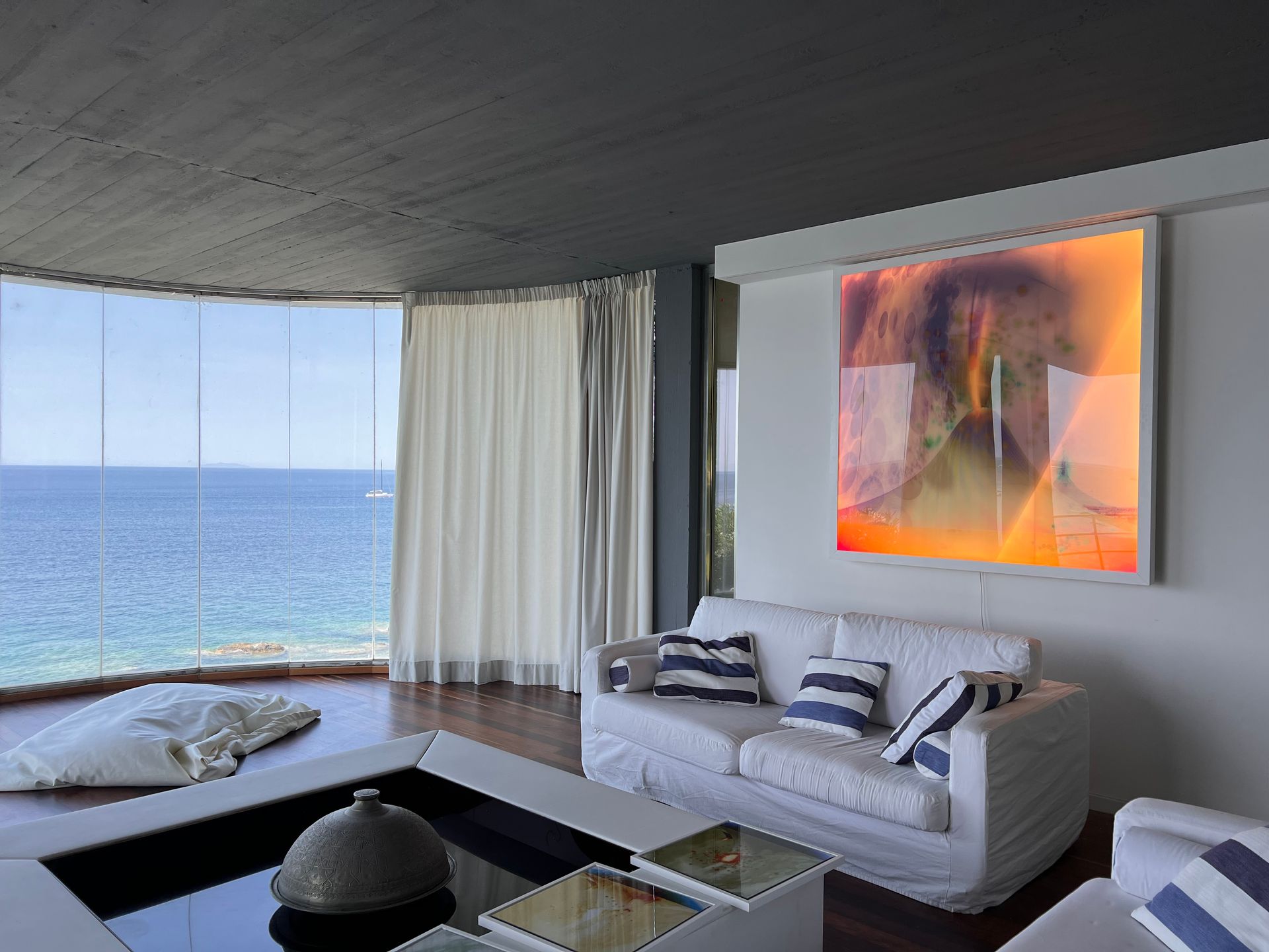 Un soggiorno moderno con divano bianco, soffitto scuro, ampia finestra con vista sul mare e vivaci opere d'arte alle pareti.